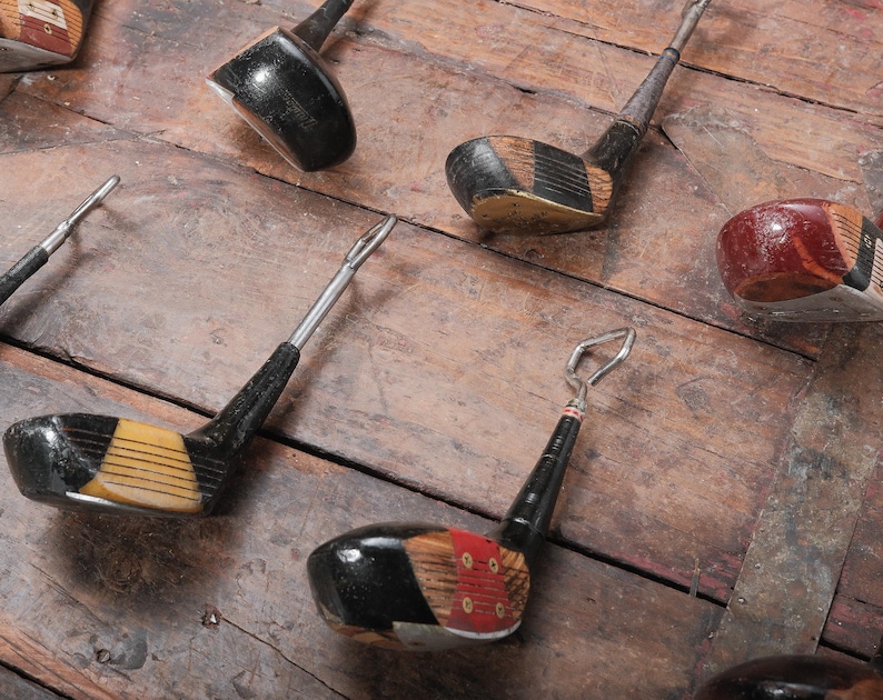 May include: A collection of vintage golf club bottle openers. The openers feature black, yellow, and red accents. The openers are arranged on a weathered wooden surface.