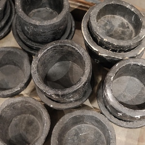 May include: A collection of dark gray, stone-like bowls and saucers. The bowls are cylindrical with a slightly textured surface, sitting on matching round saucers. The arrangement suggests a set of serving pieces or decorative items.