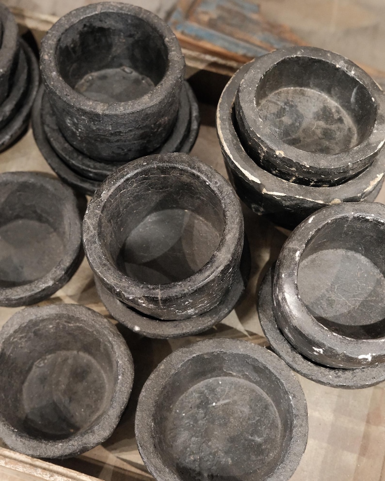 May include: A collection of dark gray, stone-like bowls and saucers. The bowls are cylindrical with a slightly textured surface, sitting on matching round saucers. The arrangement suggests a set of serving pieces or decorative items.