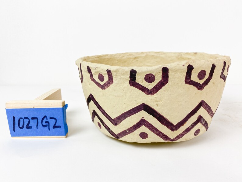 K&ouml;nnte beinhalten: Eine kleine, cremefarbene Schale mit einem geometrischen Muster aus braunen Zickzacklinien und Punkten. Die Schale ist aus Papiermach&eacute; gefertigt und hat eine raue Textur.