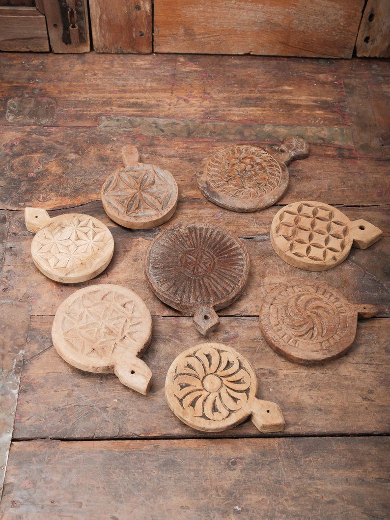 May include: A collection of eight round, wooden, hand-carved butter molds with various floral and geometric designs. The molds are in shades of light and dark brown, with handles for easy use. They are arranged on a rustic wooden surface.