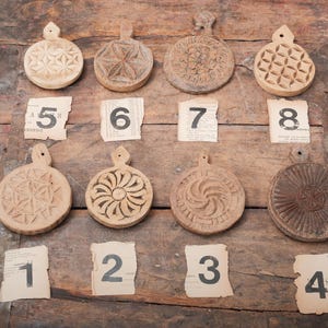 May include: A collection of eight antique wooden butter molds with various carved designs, including floral and geometric patterns. Each mold has a handle and is displayed on a rustic wooden surface with numbered paper tags.