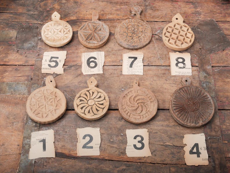 May include: A collection of eight antique wooden butter molds with various carved designs, including floral and geometric patterns. Each mold has a handle and is displayed on a rustic wooden surface with numbered paper tags.