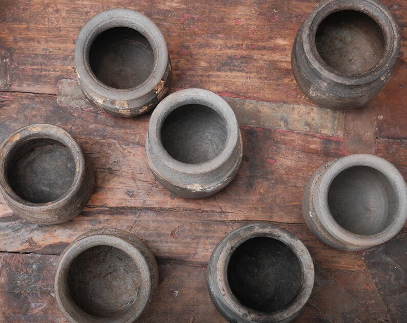 Peut inclure: Vue a&eacute;rienne de sept petits pots en c&eacute;ramique grise, us&eacute;s, avec de larges ouvertures. Les pots sont dispos&eacute;s sur une surface en bois rustique et sombre. Ils pr&eacute;sentent une texture rugueuse et des signes d'&acirc;ge.