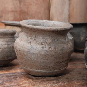 Peut inclure: Plusieurs petits pots en poterie beige, d'aspect vieilli. Les pots ont un corps arrondi et un bord large l&eacute;g&egrave;rement &eacute;vas&eacute;. La surface de la poterie pr&eacute;sente un aspect textur&eacute;. D'autres pots similaires et une surface en bois sont en arri&egrave;re-plan.