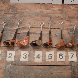 May include: A collection of eight vintage golf club bottle openers. Each opener features a wooden club head with a metal opener and a black shaft. Numbered tags from 1 to 8 are placed below the openers on a wooden surface.