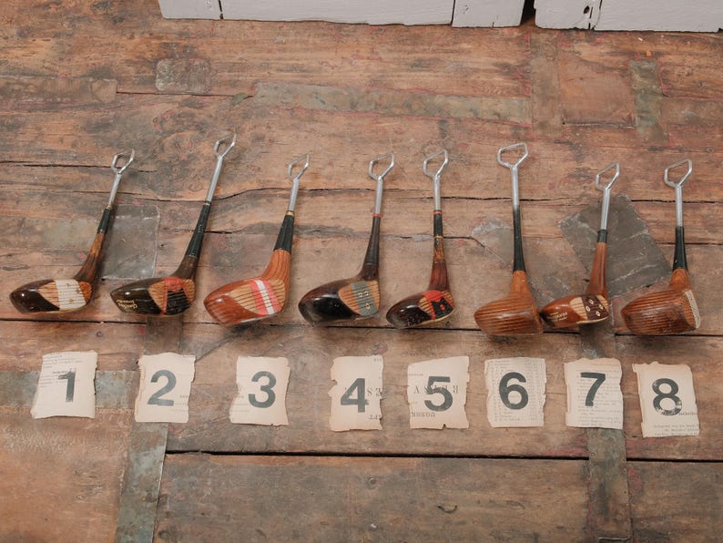 May include: A collection of eight vintage golf club bottle openers. Each opener features a wooden club head with a metal opener and a black shaft. Numbered tags from 1 to 8 are placed below the openers on a wooden surface.
