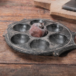 May include: A circular black cast iron pan featuring six rounded indentations arranged in a ring around a central cavity. A single, textured, brownish-red spherical object is positioned within the center of the pan, resting inside the middle well.