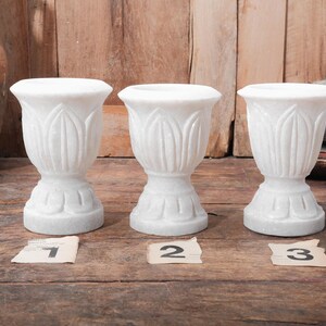 Puede incluir: Tres jarrones decorativos de m&aacute;rmol blanco con un dise&ntilde;o similar a un tulip&aacute;n. Cada jarr&oacute;n tiene una base de pedestal y una parte superior acampanada. Los jarrones est&aacute;n numerados 1, 2 y 3 en peque&ntilde;as etiquetas de papel. El fondo es de madera.