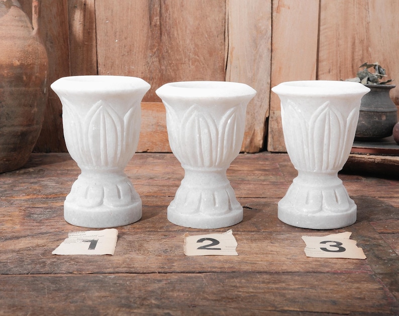 Puede incluir: Tres jarrones decorativos de m&aacute;rmol blanco con un dise&ntilde;o similar a un tulip&aacute;n. Cada jarr&oacute;n tiene una base de pedestal y una parte superior acampanada. Los jarrones est&aacute;n numerados 1, 2 y 3 en peque&ntilde;as etiquetas de papel. El fondo es de madera.