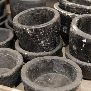 May include: A collection of dark gray stone bowls and mortar-like vessels. The bowls have a rough, textured surface with white veining. The vessels vary in size and shape, some with a cylindrical form and others with a more shallow, rounded design.