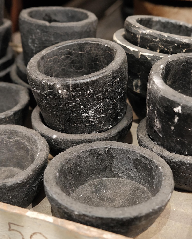 May include: A collection of dark gray stone bowls and mortar-like vessels. The bowls have a rough, textured surface with white veining. The vessels vary in size and shape, some with a cylindrical form and others with a more shallow, rounded design.