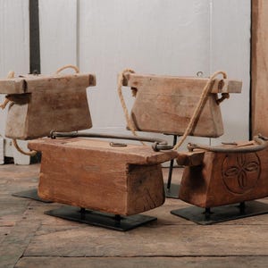 May include: Four rectangular wooden blocks mounted on black metal stands. Two blocks feature thick rope handles, while the other two have curved metal handles. One block displays a carved circular floral motif on its side, and all show weathered textures.