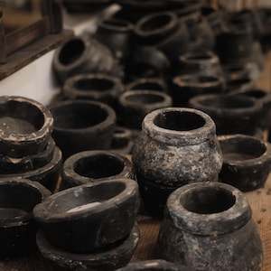 May include: A collection of dark gray, weathered stone bowls and small pots. The vessels vary in size and shape, with rounded and cylindrical forms. The objects are arranged on a wooden surface, creating a rustic, antique aesthetic.