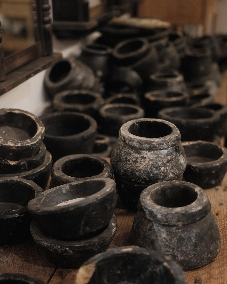 May include: A collection of dark gray, weathered stone bowls and small pots. The vessels vary in size and shape, with rounded and cylindrical forms. The objects are arranged on a wooden surface, creating a rustic, antique aesthetic.