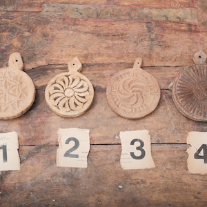 May include: Four antique wooden butter molds with intricate carved designs, each with a handle. Numbers 1 through 4 are printed on torn paper scraps below the molds. The molds are arranged on a rustic wooden surface.