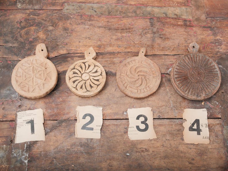 May include: Four antique wooden butter molds with intricate carved designs, each with a handle. Numbers 1 through 4 are printed on torn paper scraps below the molds. The molds are arranged on a rustic wooden surface.