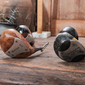 May include: Vintage golf clubs with wooden heads and metal faces, including a "MacGregor" driver and a "Northwestern Golf Co." club. The clubs are numbered and show signs of wear, resting on a wooden surface.