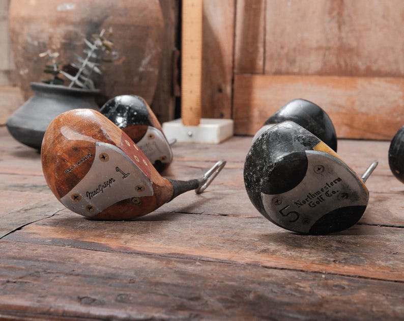 May include: Vintage golf clubs with wooden heads and metal faces, including a "MacGregor" driver and a "Northwestern Golf Co." club. The clubs are numbered and show signs of wear, resting on a wooden surface.