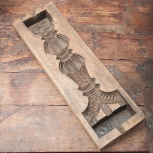 May include: A rectangular, wooden mold with a carved, ornate design. The design features a central, vertical column with decorative elements, including what appears to be a crown and leaf-like details. The wood has a weathered, aged appearance.