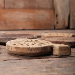 May include: A close-up of a wooden, round trivet with a carved floral design. The trivet has a handle and is light brown in color. Other similar trivets are in the background, all on a wooden surface.