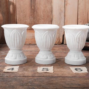 Puede incluir: Tres urnas de m&aacute;rmol blanco con un dise&ntilde;o en forma de tulip&aacute;n. Las urnas tienen una parte superior ancha y una base detallada. Las urnas se exhiben sobre una superficie de madera. Cada urna tiene una peque&ntilde;a etiqueta de papel con un n&uacute;mero.