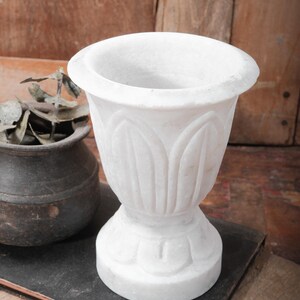 Puede incluir: Una maceta de m&aacute;rmol blanco con un dise&ntilde;o floral detallado. La maceta tiene una parte superior ancha y redondeada y una base de pedestal. El fondo presenta una superficie de madera y una peque&ntilde;a maceta de color oscuro.
