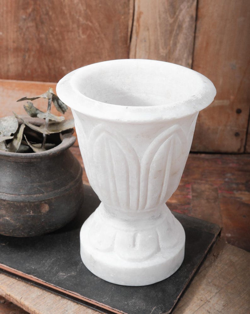 Puede incluir: Una maceta de m&aacute;rmol blanco con un dise&ntilde;o floral detallado. La maceta tiene una parte superior ancha y redondeada y una base de pedestal. El fondo presenta una superficie de madera y una peque&ntilde;a maceta de color oscuro.