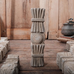 May include: A collection of weathered, light gray architectural elements. A tall, stacked column with decorative details stands in the center, flanked by smaller, rectangular pieces. The background features a wooden surface and a wooden wall.
