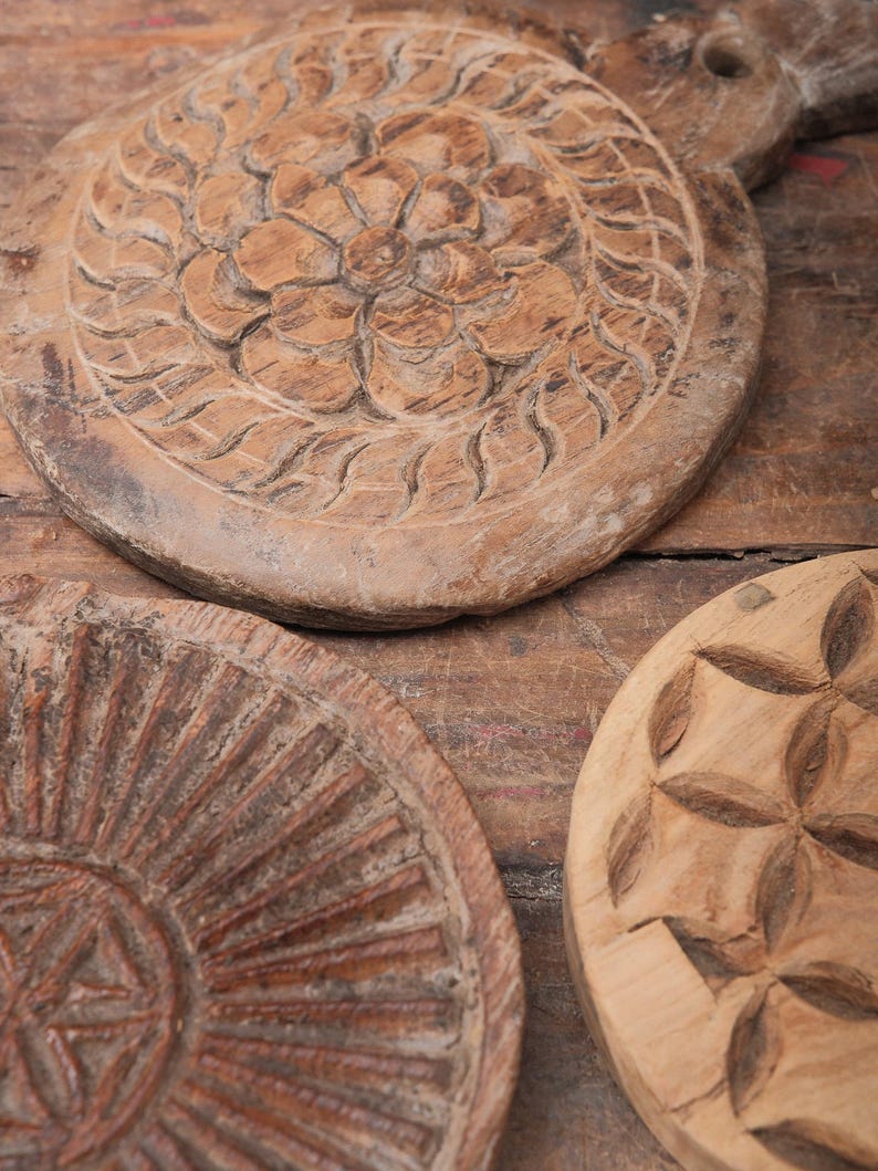 May include: Close-up of several antique wooden serving boards with carved floral and geometric designs. The boards are various sizes and shapes, with a rustic, weathered appearance. The wood has a warm, brown tone.