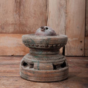 May include: A weathered, antique wooden decorative object with a round top and a base. The object has a dark, aged appearance with visible wear and tear. The top features a small, round, dark-colored element. The object is likely a decorative piece.