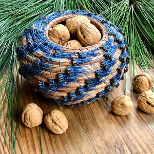 May include: A handcrafted woven basket with blue and natural fibers, filled with walnuts. The basket is round and has a textured design. Several walnuts are scattered around the basket on a wooden surface, with green pine needles in the background.