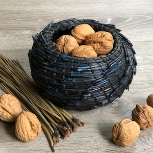 May include: A woven, dark blue basket filled with walnuts. Several walnuts are scattered around the basket. A bundle of pine needles rests beside the basket, creating a rustic, natural aesthetic.