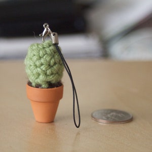 May include: A small green crocheted cactus plant in a terracotta pot with a black cord attached. The cactus is a keychain charm.