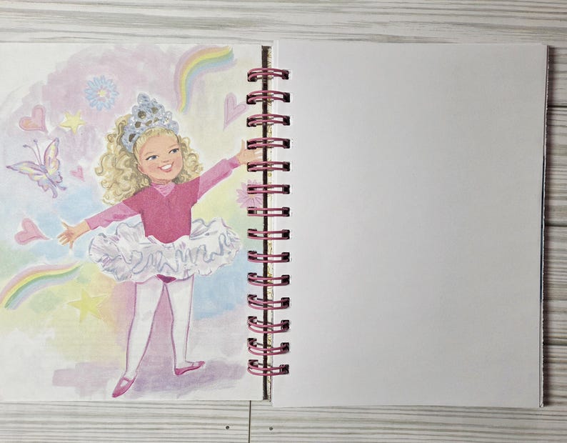 May include: A spiral-bound notebook with a colorful illustration of a young girl in a pink leotard and tutu, with a crown, surrounded by hearts, butterflies, and rainbows. The notebook has a blank page next to the illustration.