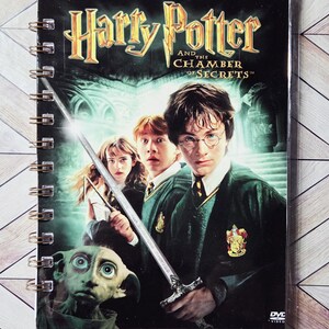 May include: A spiral-bound notebook featuring the DVD cover art for "Harry Potter and the Chamber of Secrets." The cover displays Harry, Ron, and Hermione, along with Dobby and a sword. The title is in gold lettering against a dark background.