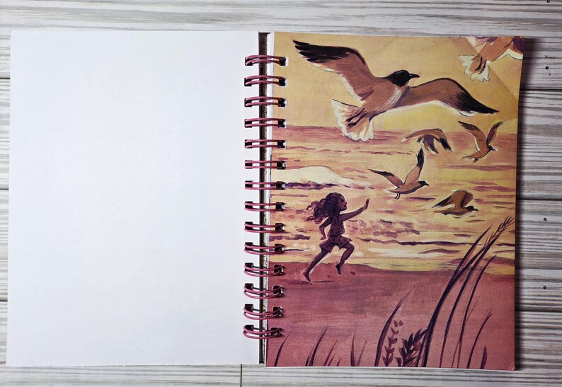 May include: A spiral-bound notebook with a blank white page and a colorful illustration. The illustration depicts a child running on a beach with seagulls flying overhead. The artwork is in shades of brown, orange, and yellow.