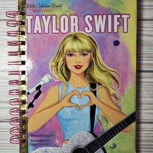 May include: A spiral-bound notebook featuring a colorful illustration of Taylor Swift. The cover art depicts a blonde woman with blue eyes holding a guitar and forming a heart shape with her hands. The title "Taylor Swift" is prominently displayed.