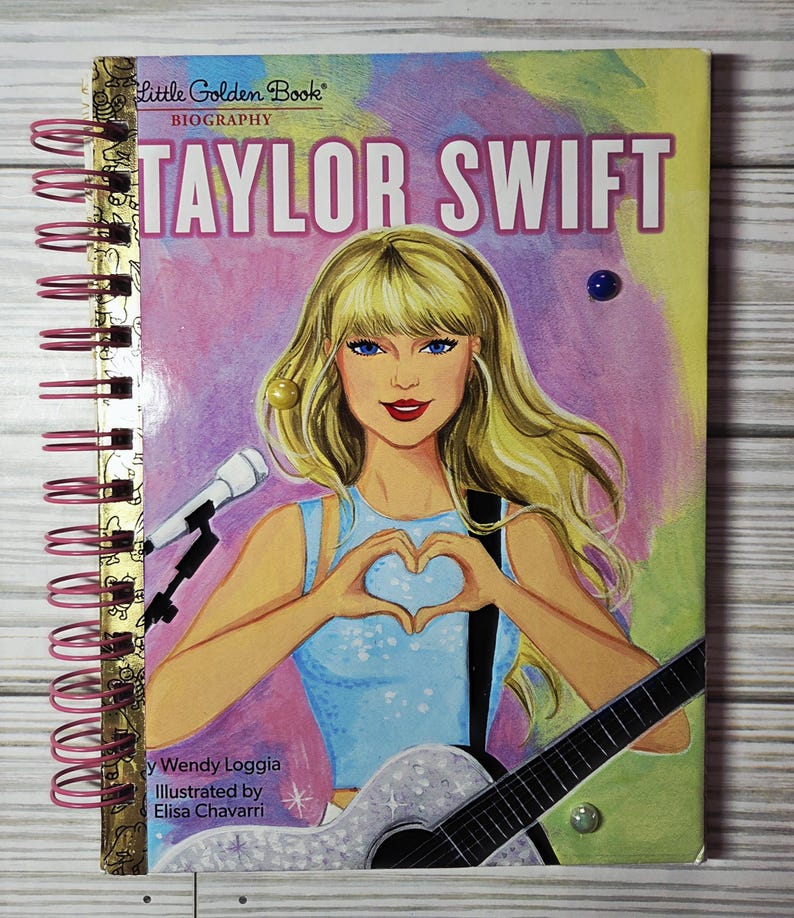 May include: A spiral-bound notebook featuring a colorful illustration of Taylor Swift. The cover art depicts a blonde woman with blue eyes holding a guitar and forming a heart shape with her hands. The title "Taylor Swift" is prominently displayed.