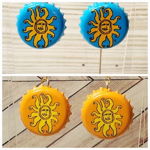 May include: A pair of earrings made from bottle caps with a yellow sun design on a blue and orange background. The earrings have gold hooks.