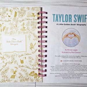 May include: Open book with a spiral binding, featuring a title page that reads "TAYLOR SWIFT: A Little Golden Book Biography." The left page has a gold floral pattern and the words "Little Golden Books." The book is open on a light wood surface.