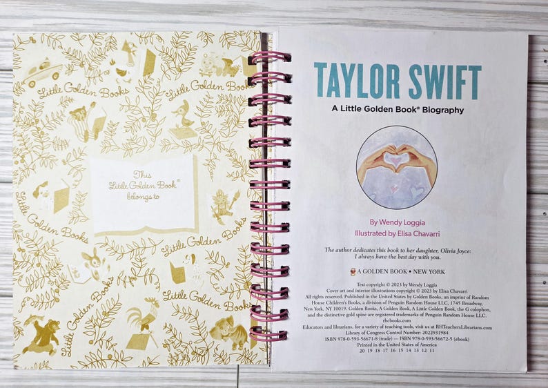 May include: Open book with a spiral binding, featuring a title page that reads "TAYLOR SWIFT: A Little Golden Book Biography." The left page has a gold floral pattern and the words "Little Golden Books." The book is open on a light wood surface.