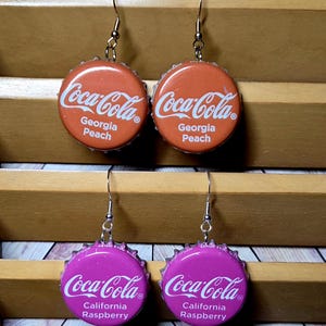 May include: Two pairs of dangle earrings made from metal bottle caps. The top pair is orange with white text reading Coca-Cola Georgia Peach. The bottom pair is purple with white text reading Coca-Cola California Raspberry. Each features silver-toned hooks.