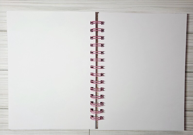 May include: Open blank notebook with white pages and a pink spiral binding. The notebook is lying open on a light gray wooden surface. The pages are clean and ready for writing or drawing.