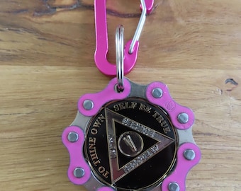 May include: A circular keychain featuring a pink bicycle chain link border surrounding a black medallion. The medallion displays a triangle with the text "TO THINE OWN SELF BE TRUE" and "UNITY SERVICE RECOVERY" with a Roman numeral V. A pink carabiner is attached.