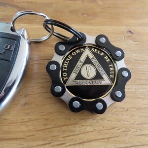 May include: A circular keychain featuring a black and silver bicycle chain link border surrounding a central medallion. The medallion displays a triangle with the text TO THINE OWN SELF BE TRUE, UNITY, SERVICE, RECOVERY, and the Roman numeral V.