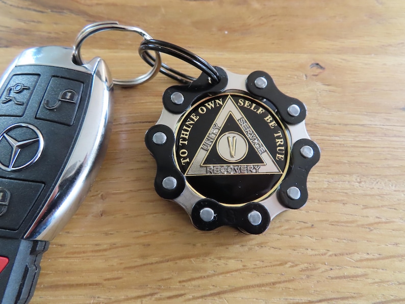 May include: A circular keychain featuring a black and silver bicycle chain link border surrounding a central medallion. The medallion displays a triangle with the text TO THINE OWN SELF BE TRUE, UNITY, SERVICE, RECOVERY, and the Roman numeral V.