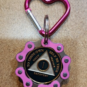 May include: A pink heart-shaped carabiner clip attached to a silver chain with a round, green and gold medallion. The medallion features a triangle with the words "To Thine Own Self Be True", "Unity", "Service", "Recovery", and the number "1" inside the triangle.