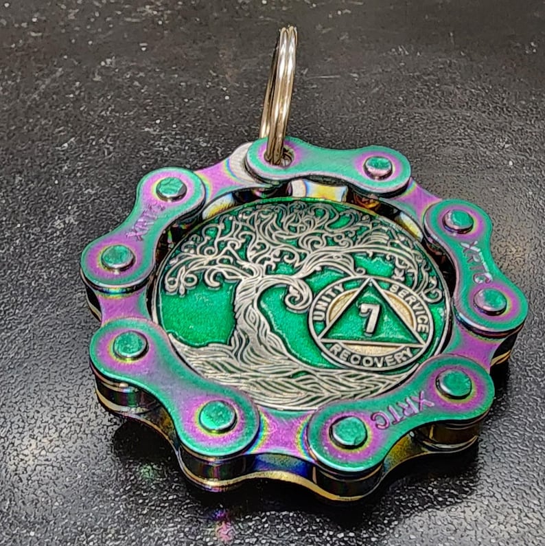 May include: A circular pendant featuring a metallic bicycle chain border with an iridescent purple and green finish. The center displays a silver tree design over a green background, with a triangle emblem containing the number 7 and the text UNITY SERVICE.