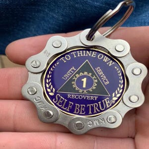 May include: A circular metal keychain featuring a silver bicycle chain link border surrounding a purple enamel medallion. The medallion displays a black triangle with the text Unity, Service, Recovery, and the number 1, with To Thine Own Self Be True.